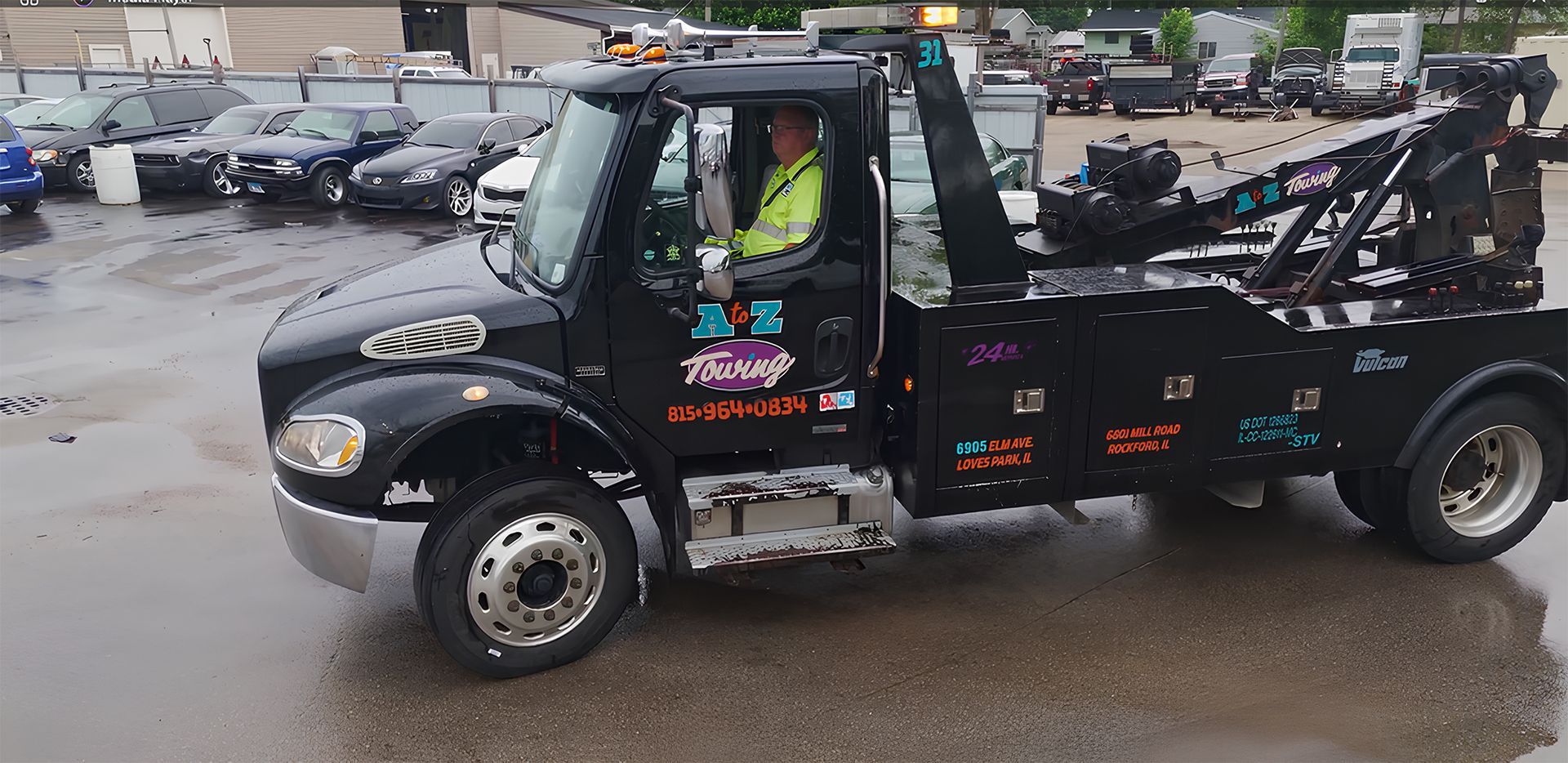 Black tow truck with a driver wearing a yellow vest, parked in a lot of cars.