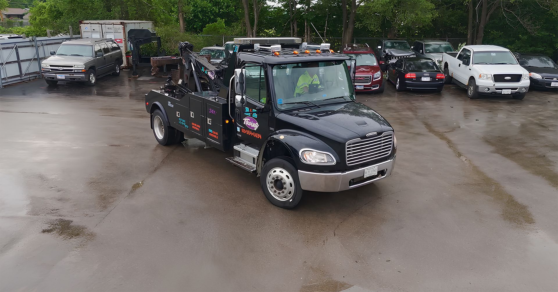 Black tow truck in a lot with parked cars. Rainy day.