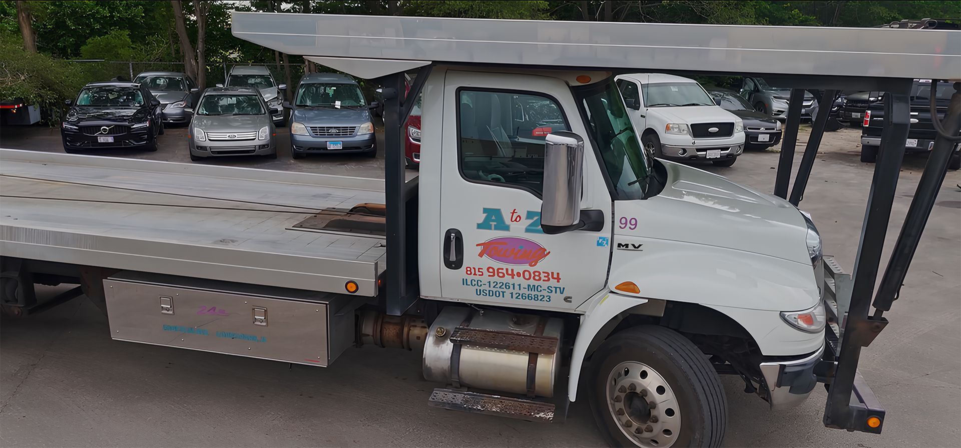 A white tow truck with a flatbed is parked near several cars. The truck has the company name 