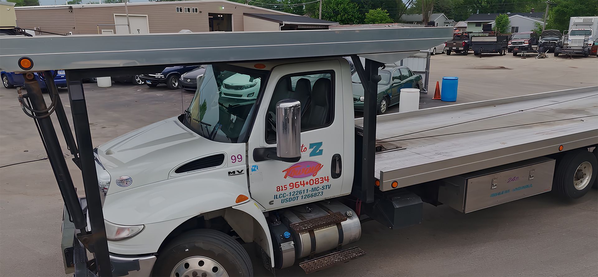 White flatbed tow truck with a driver in the cab. Parked in a lot.