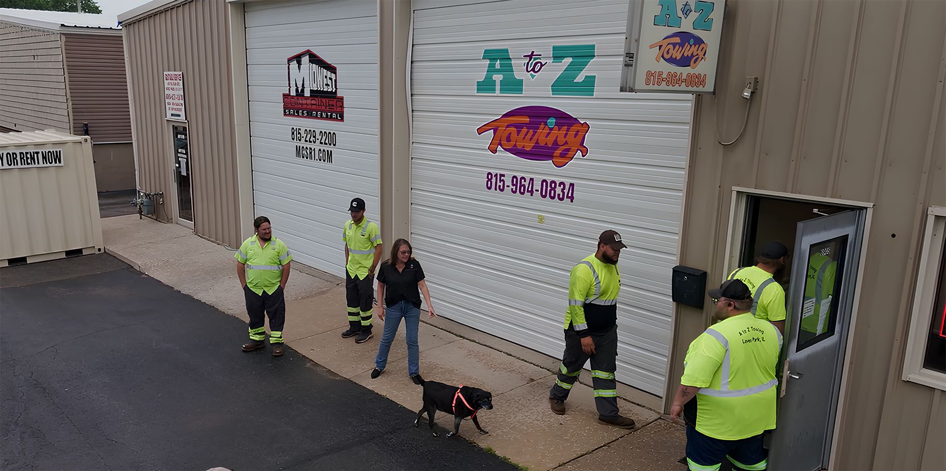 People in safety vests stand outside a building with garage doors, a dog is present.