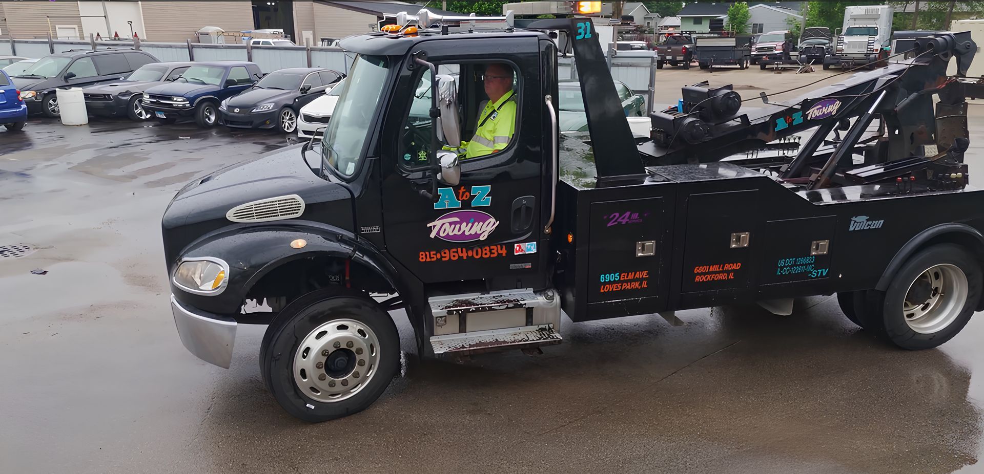 A black tow truck with a driver in a reflective vest is parked on a wet surface with other cars in the background.
