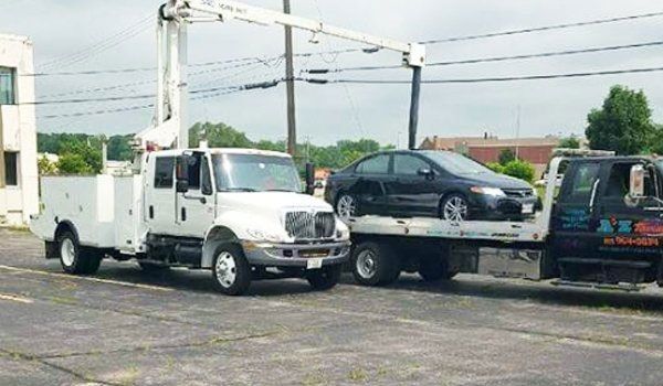 A black car being towed on a flatbed by a tow truck on a city street.