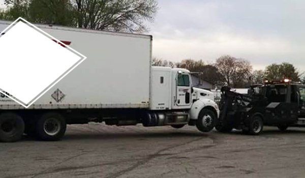 White semi-truck being towed by a black tow truck in a parking lot. Cloudy sky in the background.