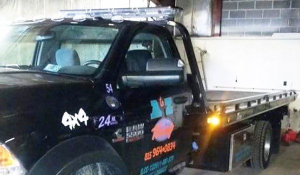 Black tow truck with a flatbed in a garage, with logo on the side that reads 