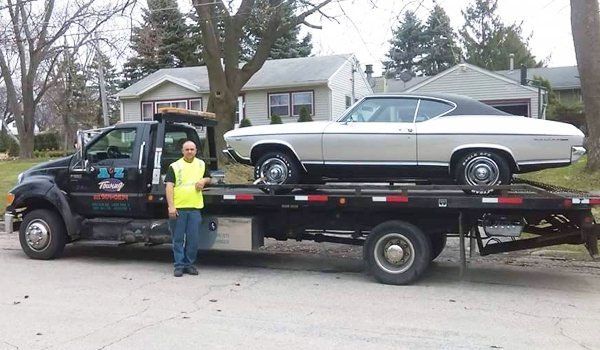 Tow truck transporting a silver classic car. A man in a safety vest stands nearby on a residential street.