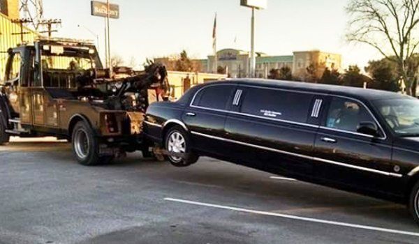 Tow truck towing a black limousine on a paved road; the car is partially lifted.