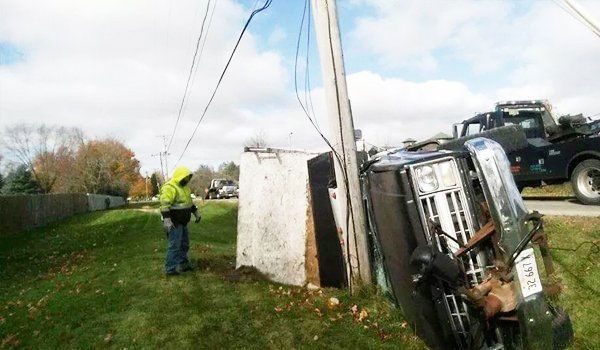 Overturned pickup truck against a utility pole. A person in a high-vis jacket assesses the scene. A tow truck is present.