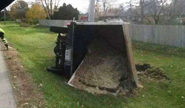 A black truck overturned on grass next to a road, bed up and filled with dirt.