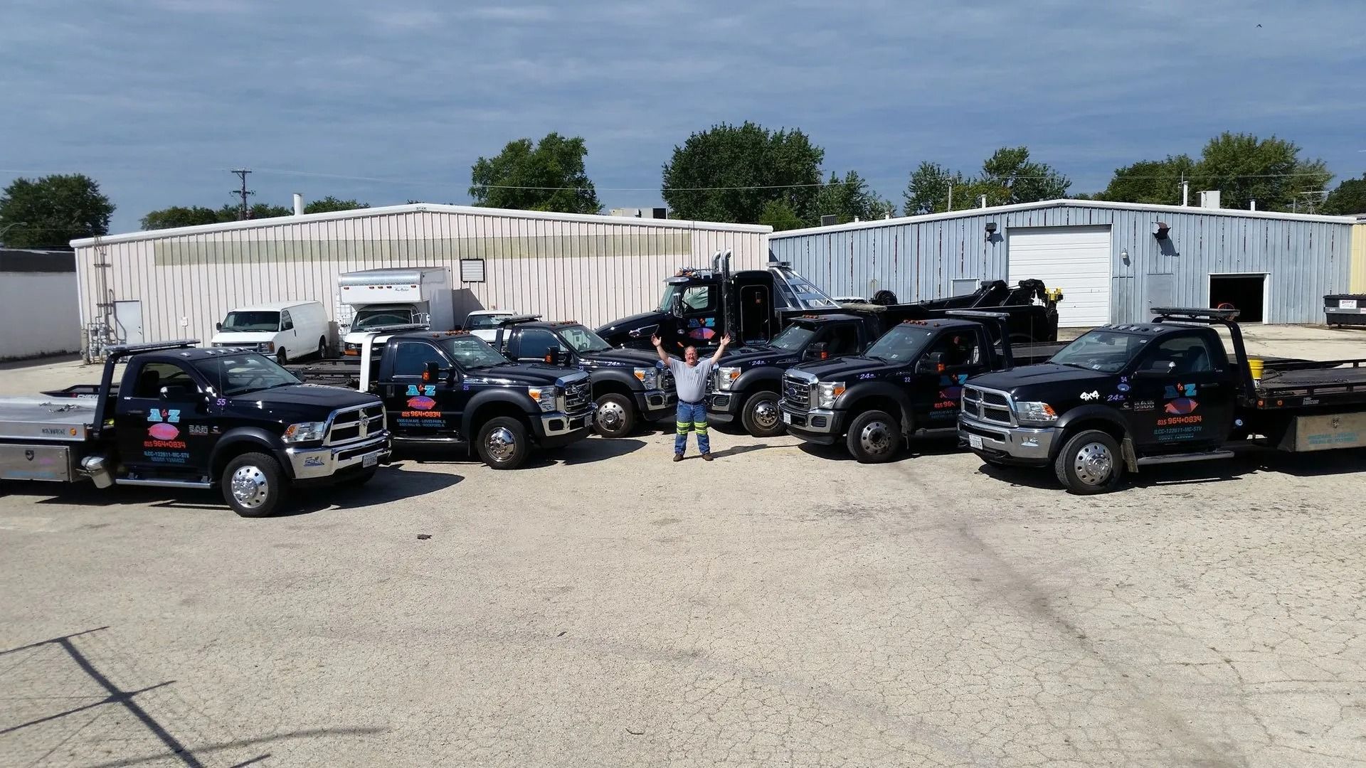 Five black tow trucks parked, man in the middle with arms raised, sunny day.