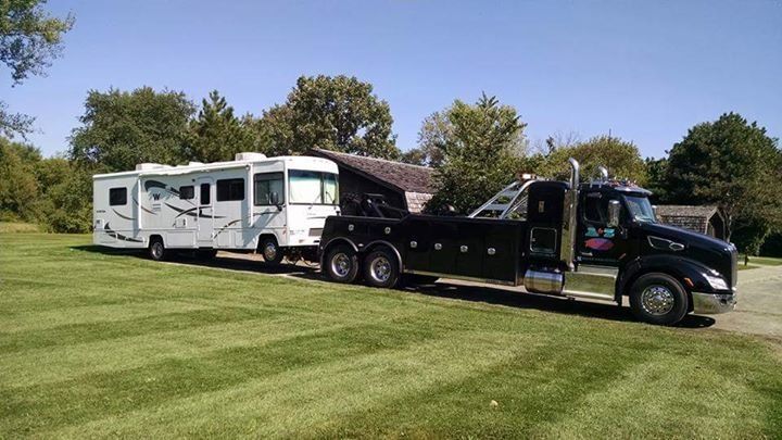 A black tow truck towing a white RV on a grassy lawn under a blue sky.