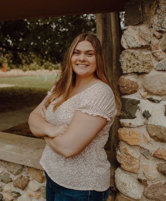 Woman with arms crossed, smiling, standing beside a stone wall. Outdoors, wearing floral top and jeans.