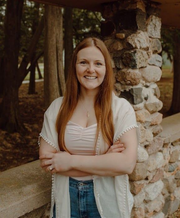 Woman with red hair and a pink top smiles while standing in front of a stone wall with crossed arms.