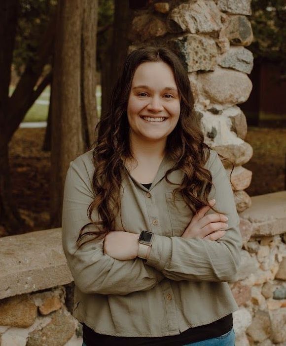 Woman with arms crossed, smiling, wearing a green shirt, standing by a stone wall outside.