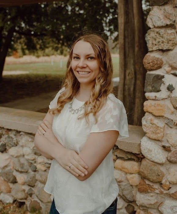 Woman with arms crossed, smiling, standing near a stone wall and wooden structure. She wears a white blouse.