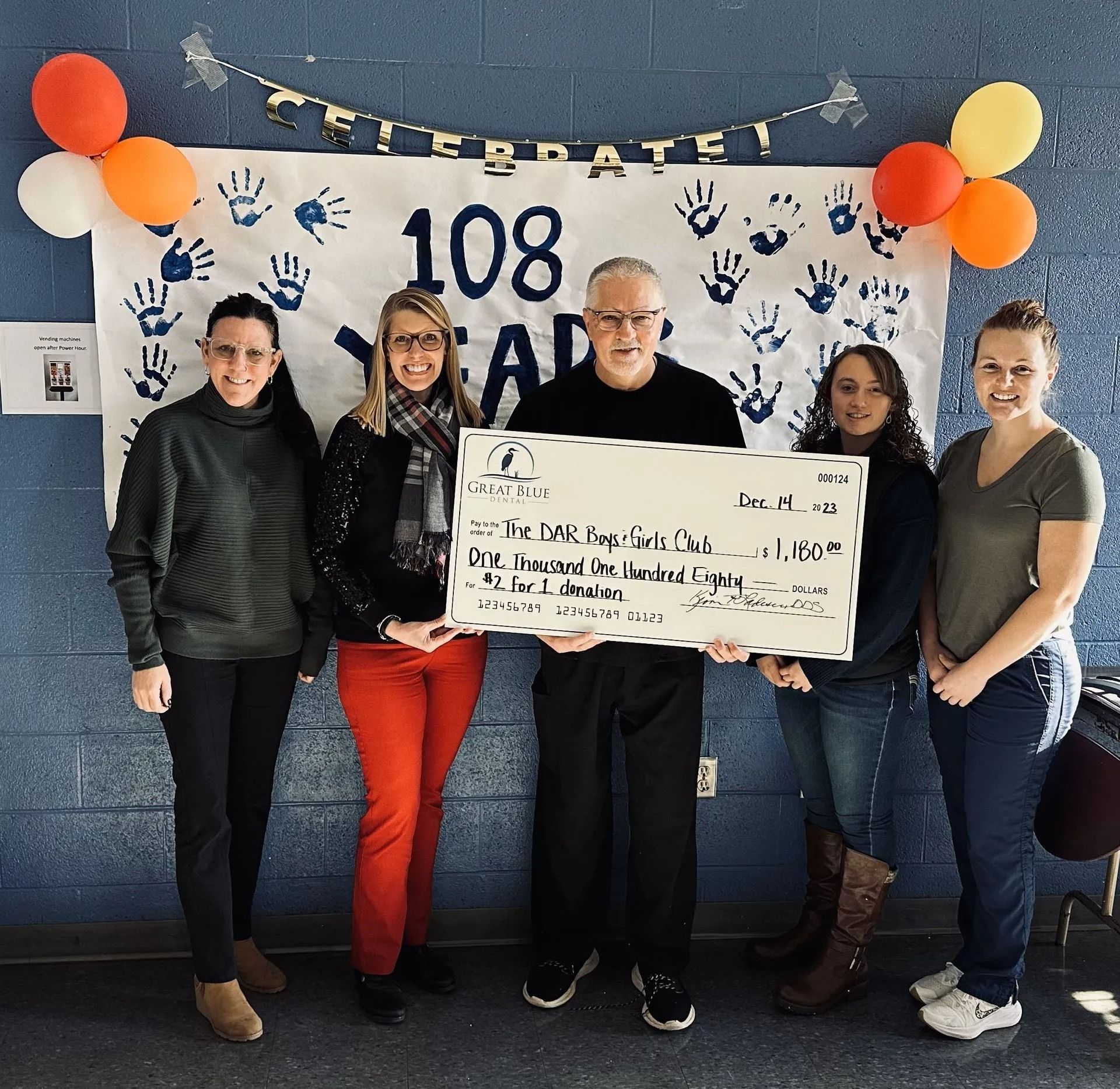 Five people holding a large check, celebrating 108 years. Banner and balloons in background.
