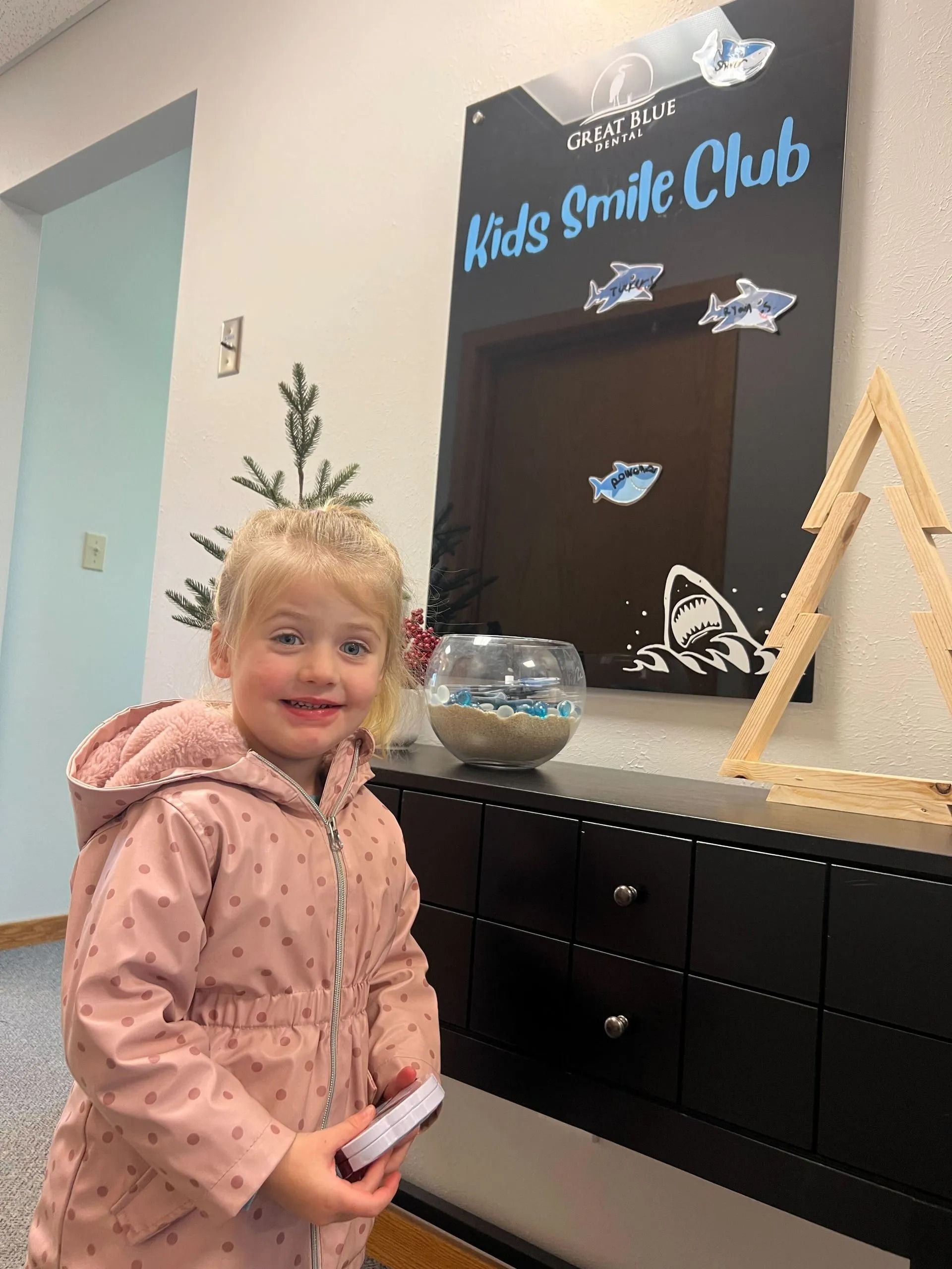 Young girl smiles near "Kids Smile Club" sign with fish decorations.