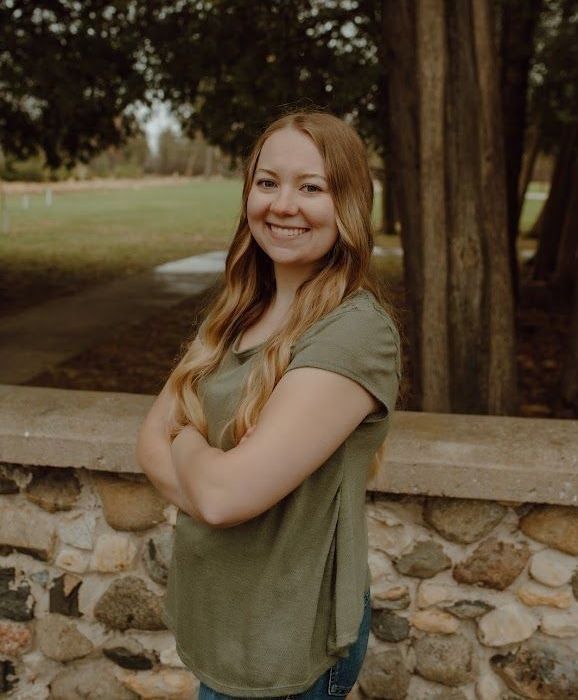 Woman with long blonde hair, smiling, arms crossed, in green shirt in front of a stone wall.