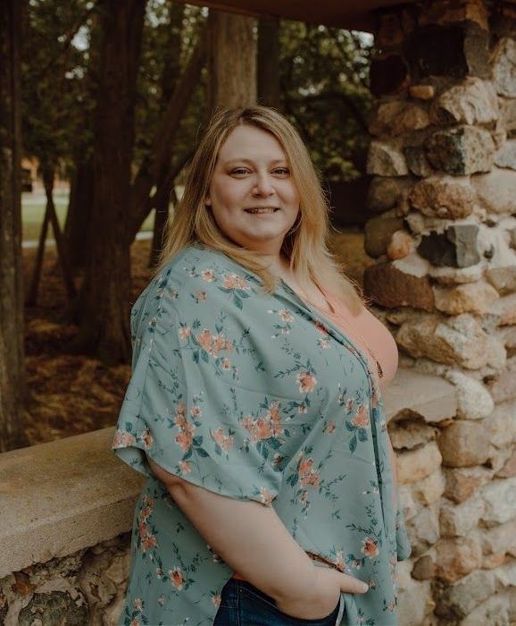 Woman with blonde hair smiles, wearing a floral kimono, standing near a stone wall outdoors.
