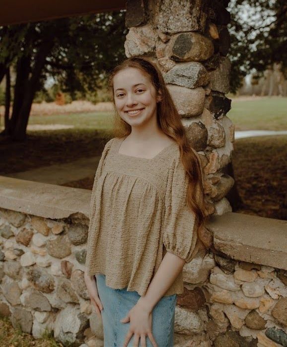 Young woman smiles, leaning against a stone wall. She wears a tan top and blue jeans, outdoors.