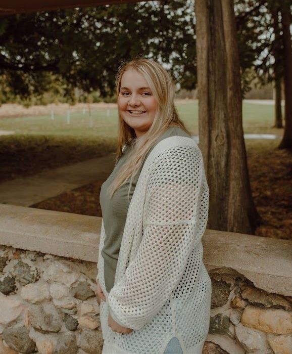 Woman with blonde hair smiles in a park, wearing a white sweater and olive green shirt.