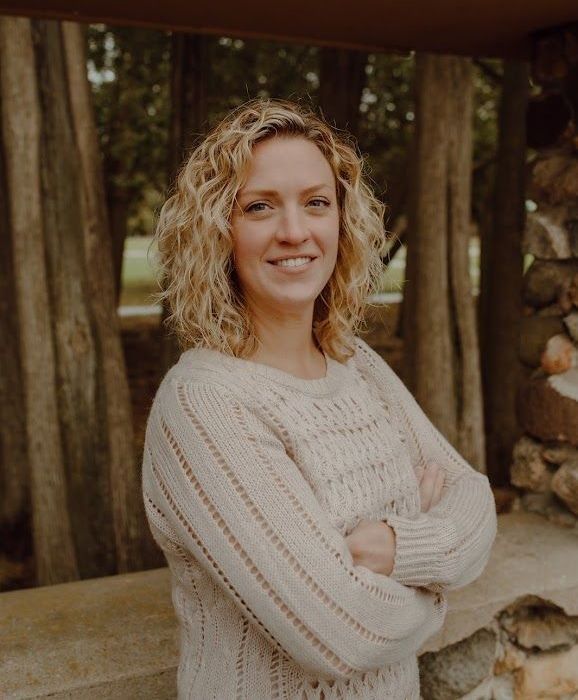 Woman with curly blonde hair and crossed arms smiles, wearing a cream sweater outdoors with trees.