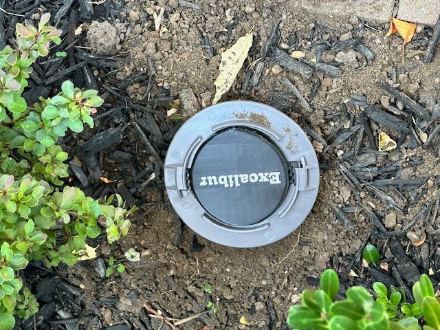 An Excalibur-branded subterranean termite bait station installed in mulch near green landscaping.