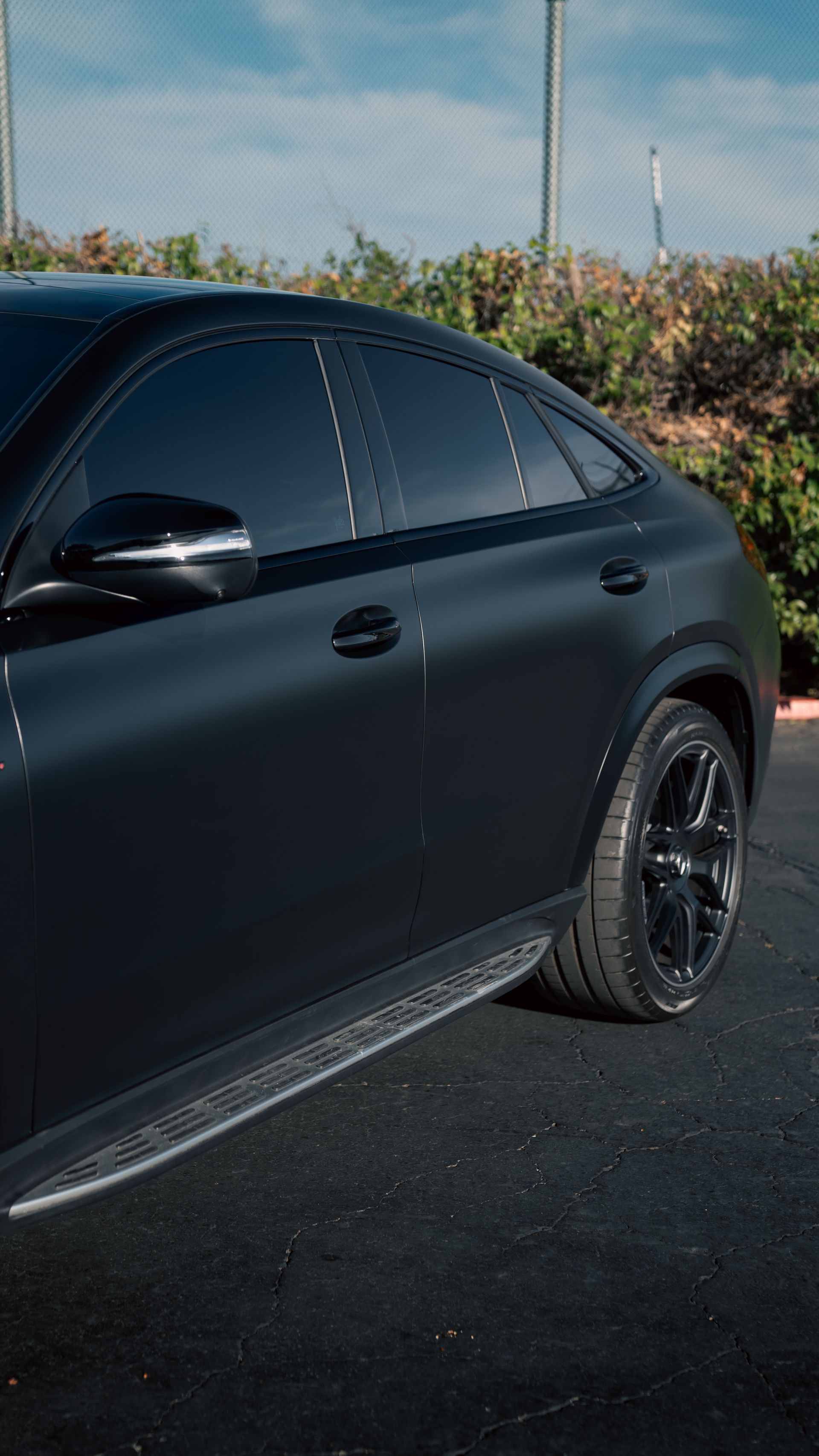 Black sedan parked outdoors on asphalt with tinted windows and black wheels, near bushes under a blue sky