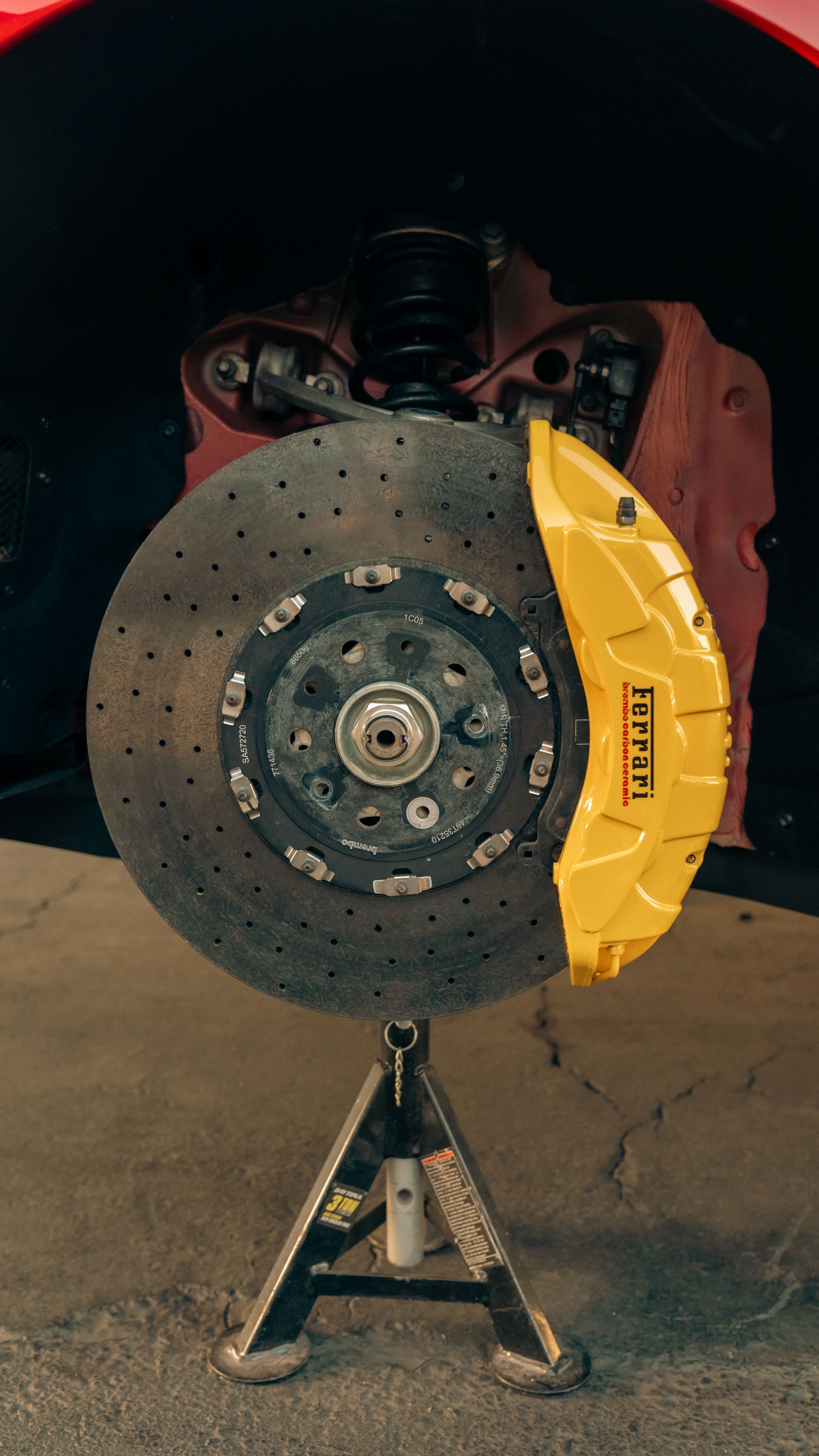 Car’s front wheel with brake rotor exposed and a yellow caliper, raised on a jack in a garage.