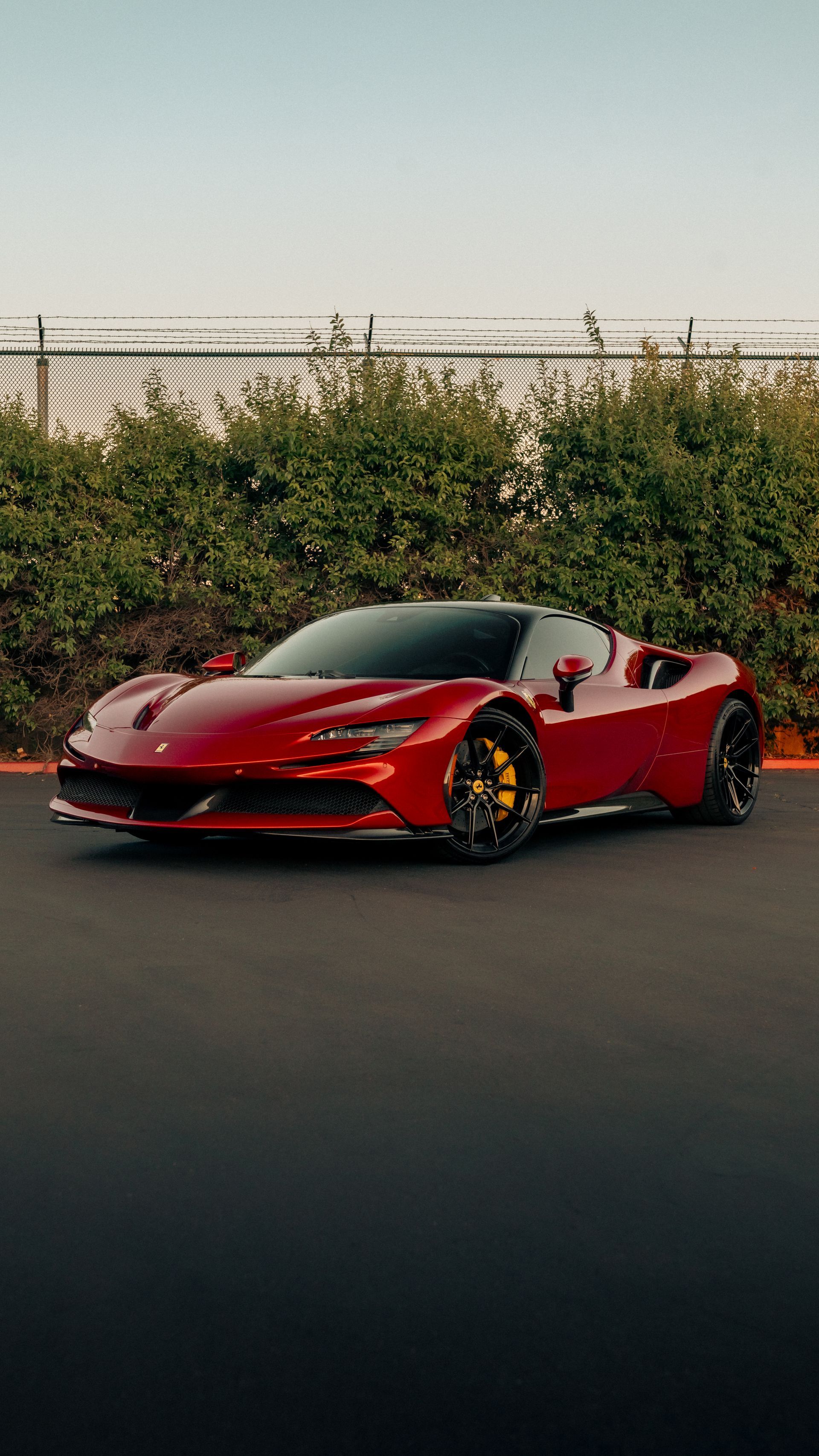 Red sports car parked on a road at sunset, with a chain-link fence and greenery behind it.