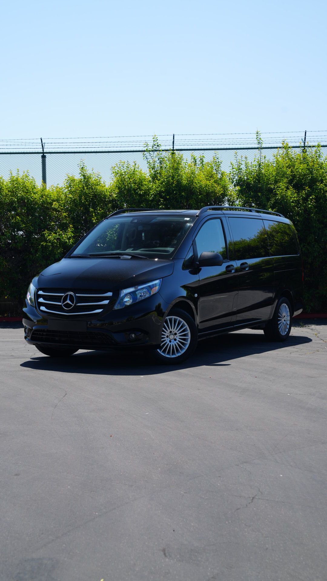 Black Mercedes van parked on a sunny paved lot beside a chain-link fence and greenery.