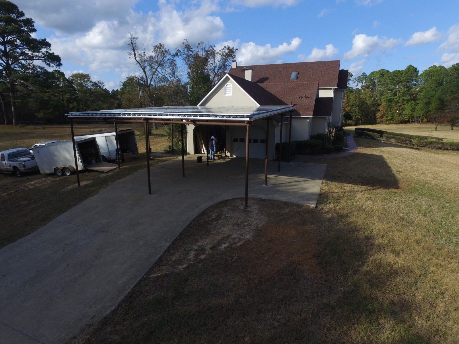 A residential house with a large metal carport in the driveway, adjacent to parked trailers on a sunny, grassy lot.