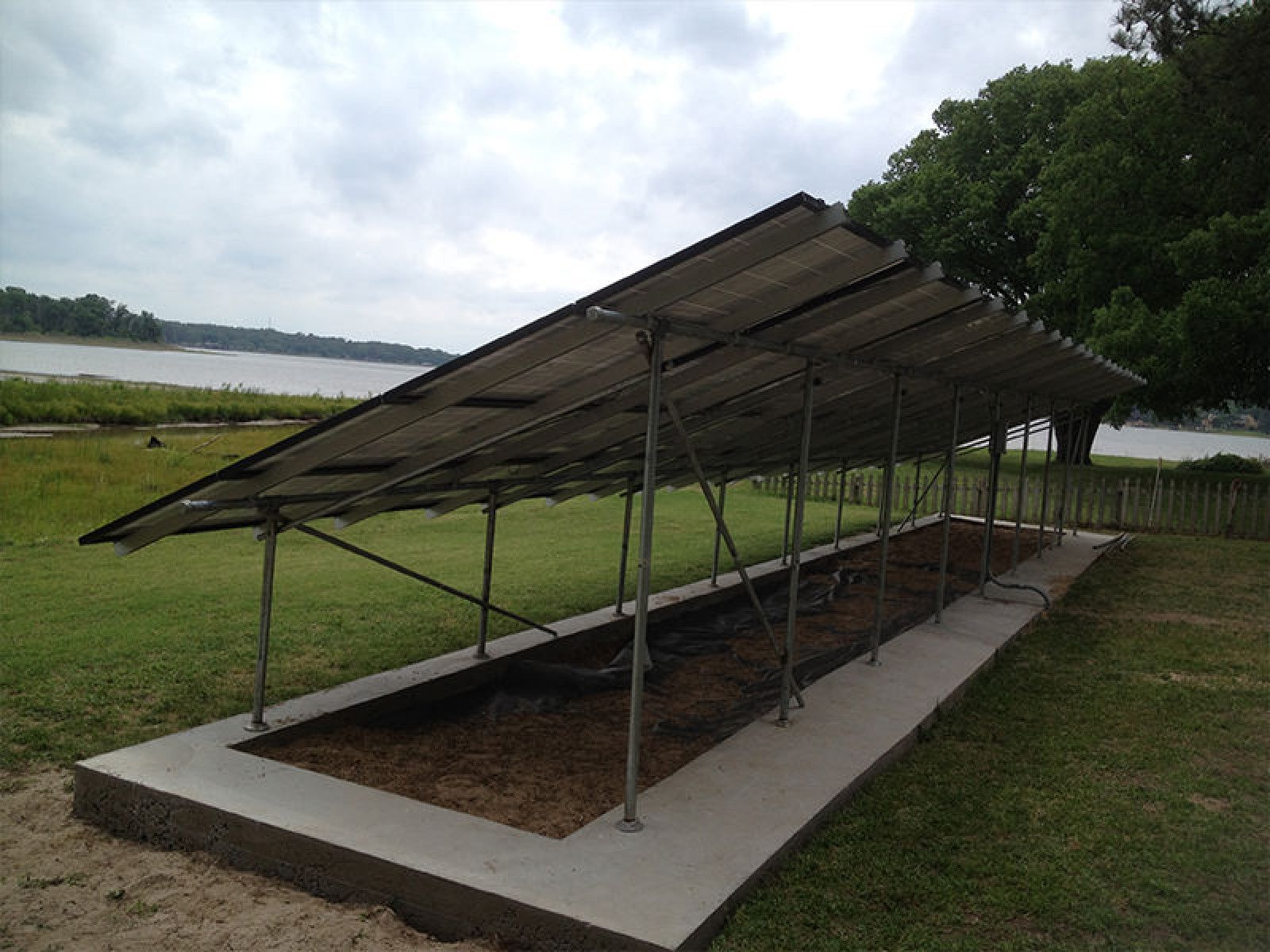 A row of solar panels mounted on a metal frame above a concrete base in a grassy, lakeside field.