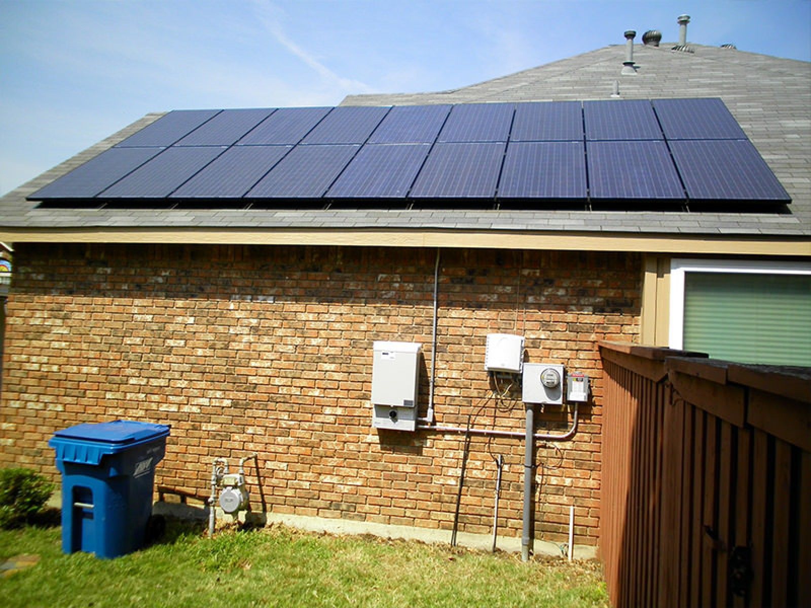 Solar panels installed on a residential roof above a brick wall with exterior electrical equipment and a blue bin.