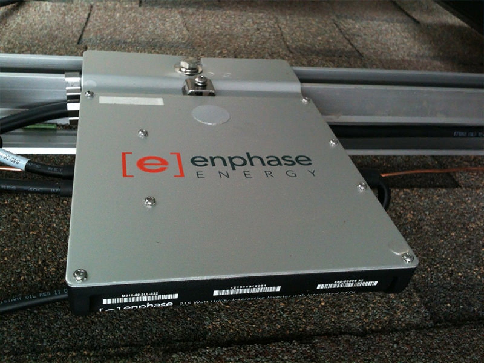 An Enphase Energy microinverter mounted on metal rails atop a dark shingled roof.