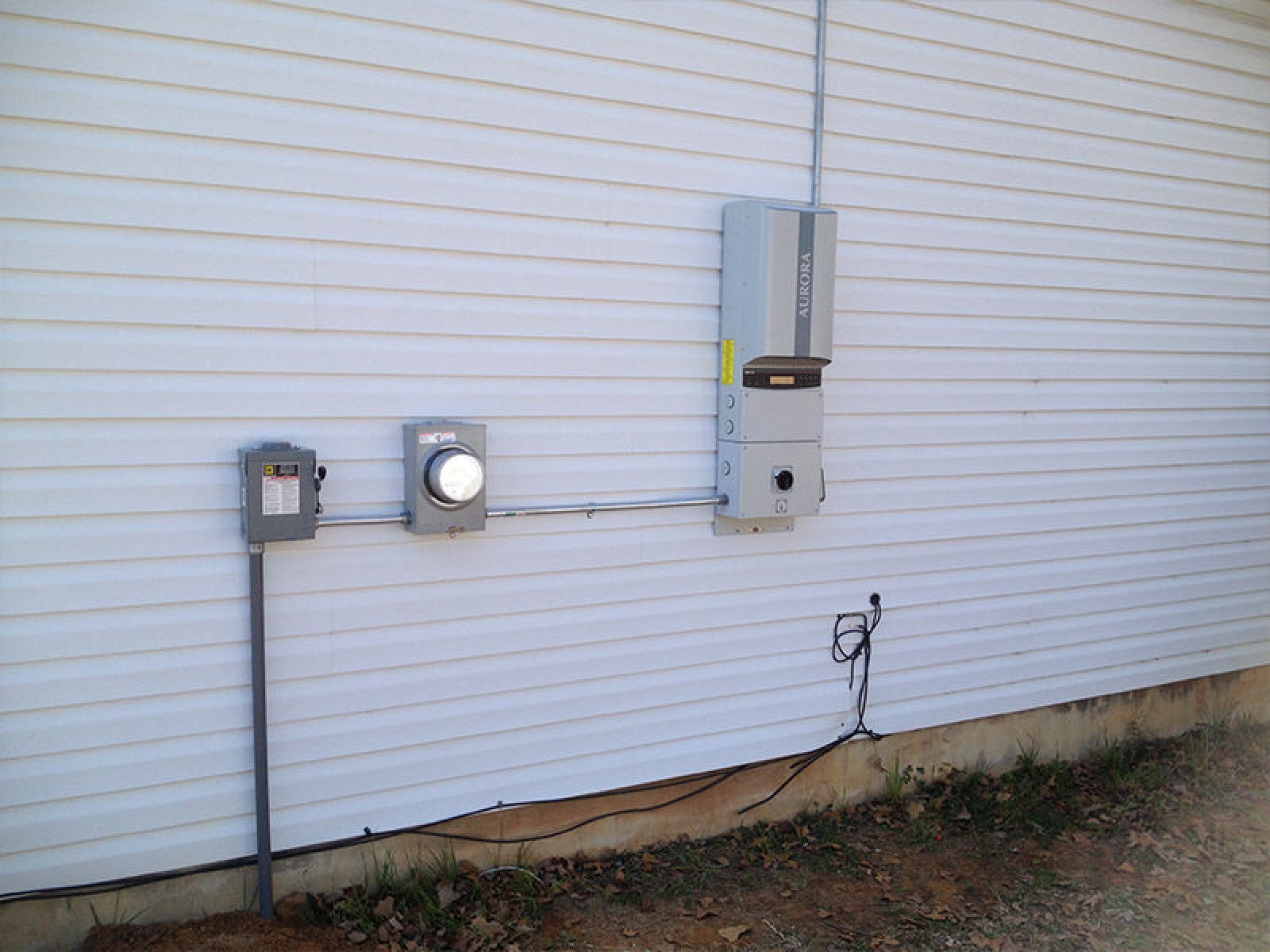 An exterior wall with gray vinyl siding featuring an electrical meter, a disconnect box, and a solar power inverter.