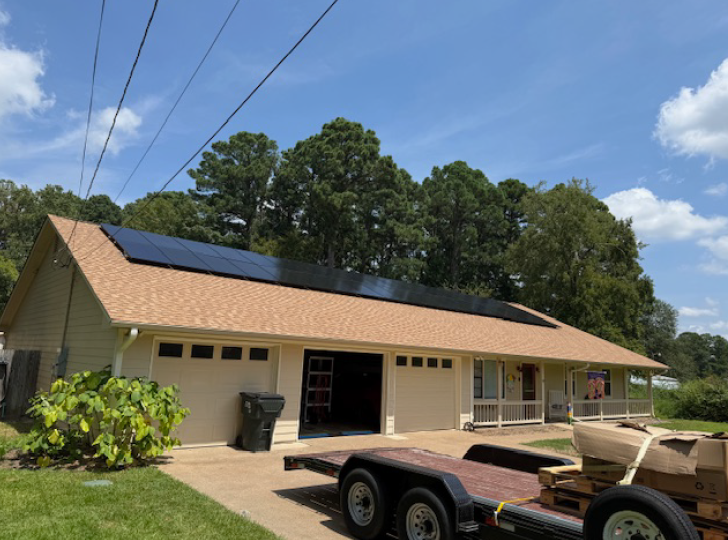 A single-story tan house with a garage and solar panels installed on the roof, viewed from a driveway with a trailer.