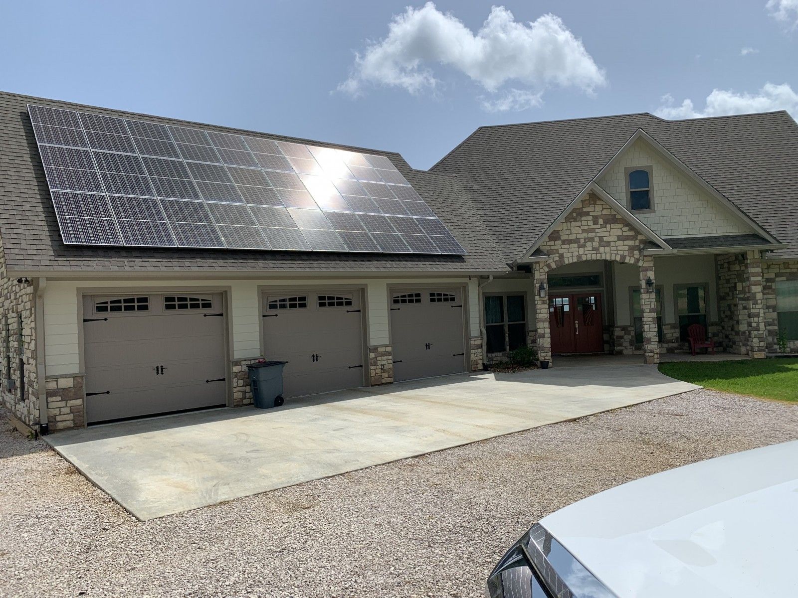 A light-colored house with a large solar panel array mounted on the roof above a three-car garage and a gravel driveway.