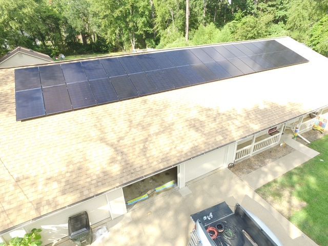An aerial view of a row of black solar panels installed on the shingled roof of a house with a surrounding lawn.