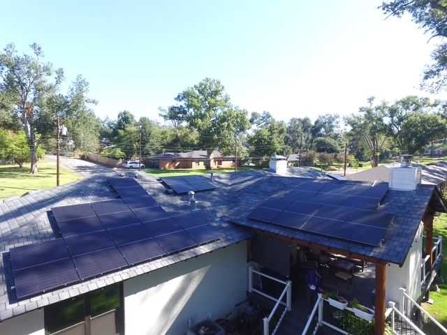 An elevated view of a residential roof covered with black solar panels, with surrounding trees and houses in the distance.
