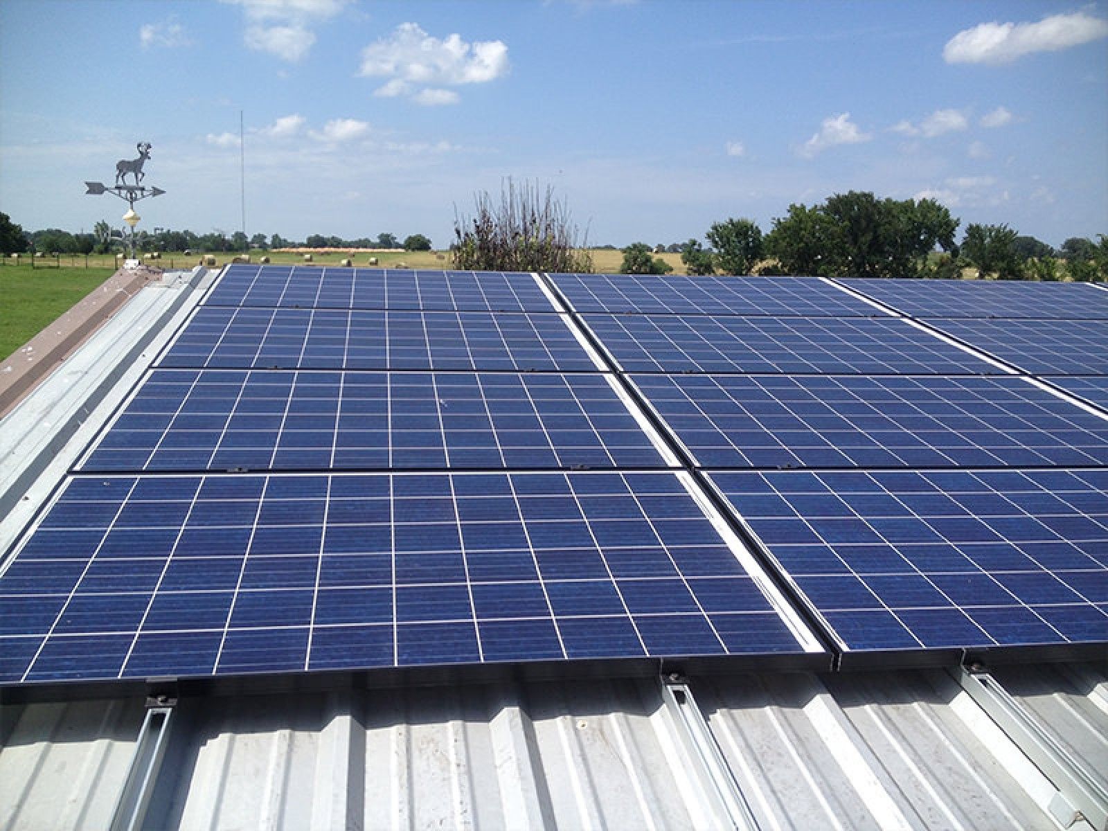 Rows of blue solar panels mounted on a corrugated metal roof under a bright, sunny sky with a weather vane in the distance.