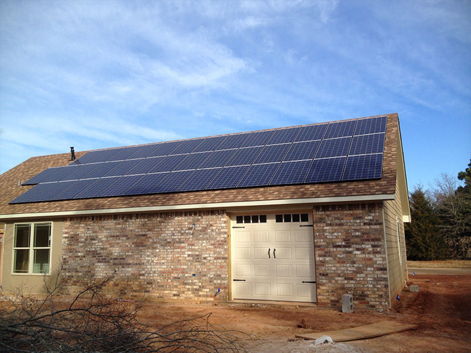 A tan brick building with a garage door and solar panels installed on the roof under a blue, cloudy sky.