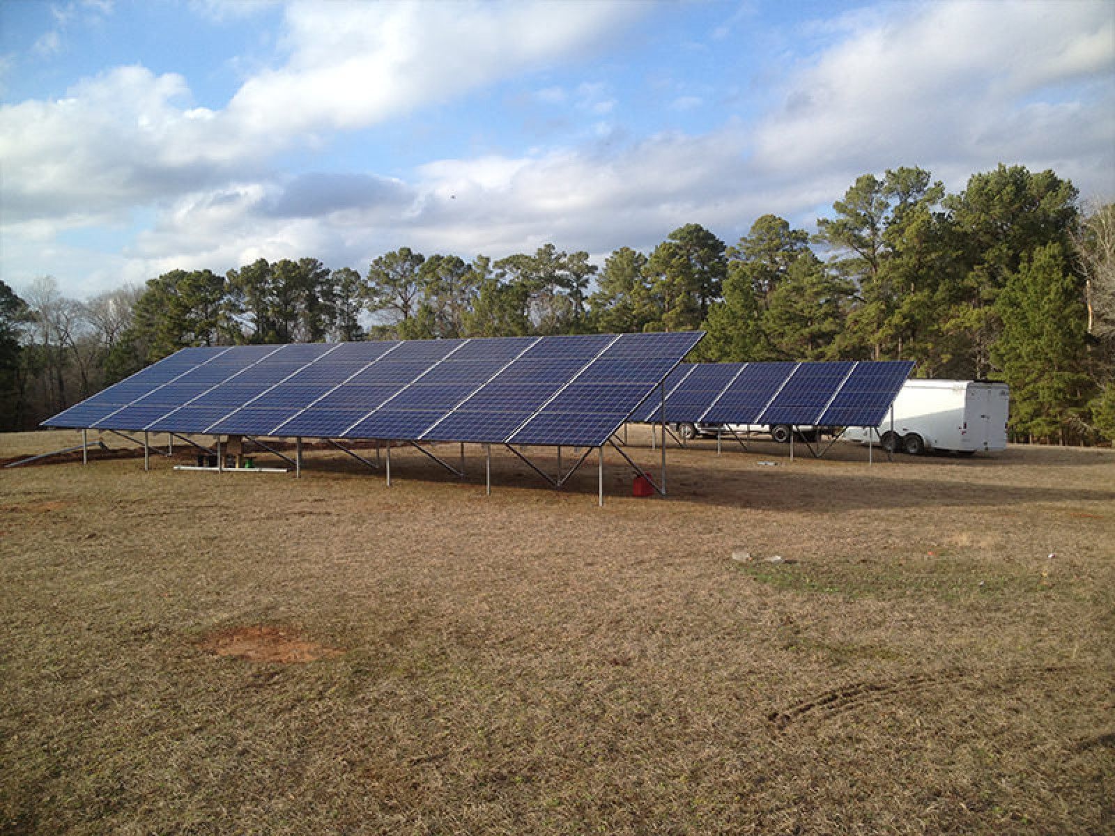 Ground-mounted solar panel array installed in a large, open field with a trailer nearby and a line of trees in the back.
