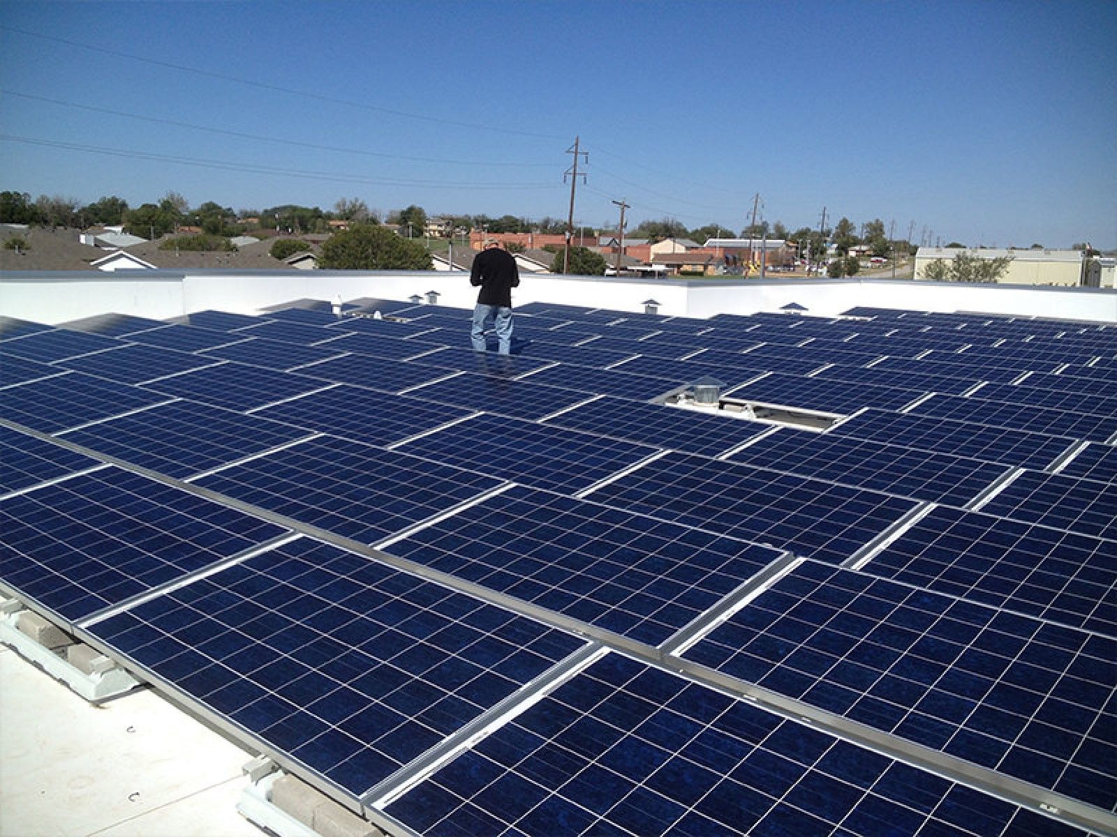 A person standing on a flat roof covered in a grid of blue solar panels under a clear blue sky.