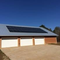 A tan garage with three white bay doors and solar panels installed on the sloped metal roof under a clear blue sky.