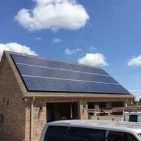 A brick garage building with a large solar panel array covering the entire roof against a blue sky with white clouds.
