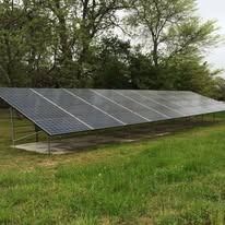A row of dark solar panels mounted on a metal frame sits in a grassy field in front of a line of trees.