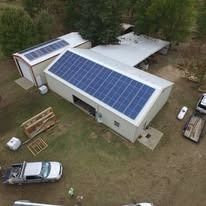 Aerial view of two white metal buildings with solar panel arrays on their roofs, situated on a grassy, wooded lot.