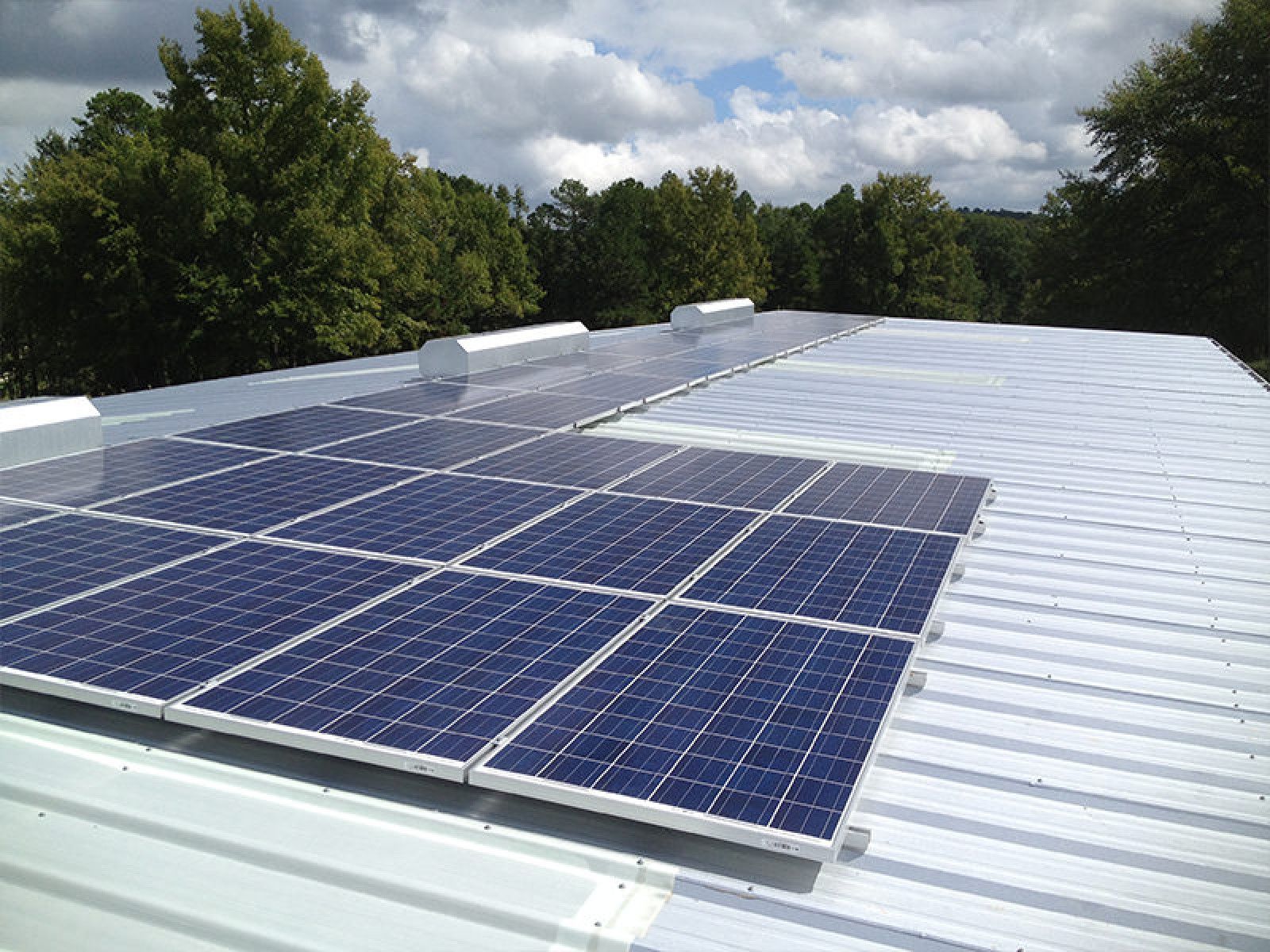 A partial array of rectangular, blue solar panels mounted on a silver, corrugated metal roof under a cloudy sky.