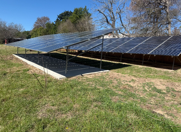 Two rows of ground-mounted solar panels installed over a concrete pad in a grassy, outdoor area with trees.